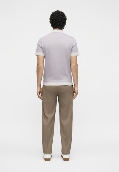 Άνδρας με σγουρά μαλλιά που φοράει ένα ανοιχτό λεβαντίνι κοντομάνικο πόλο πουκάμισο και καφέ παντελόνια, στέκεται γυρισμένος με την πλάτη προς τα εμπρός σε λευκό φόντο.