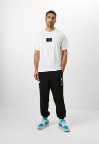 Witte katoenen t-shirt met een zwart Nike-logo, gecombineerd met zwarte joggingsbroeken met gouden accenten en het NBA-logo, afgemaakt met blauwe sneakers.