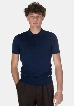 Giovane uomo con capelli ricci che indossa una polo blu navy aderente a maniche corte e pantaloni marroni scuri, in piedi con una mano in tasca.
