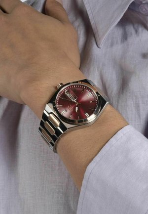 Silver and gold metal watch with a red dial, featuring a date window, stick hour markers, and a polished bracelet with alternating links.