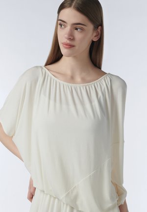 Jonge vrouw met lang bruin haar die een losse, crèmekleurige blouse met geplooide hals draagt, kijkt opzij tegen een lichte achtergrond.