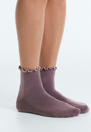Legs wearing ribbed purple ankle socks with ruffled edges, standing on a smooth white surface.