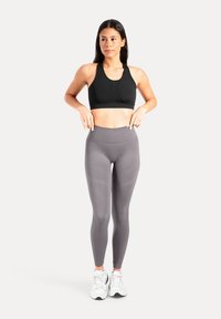 Brassière de sport noire et leggings gris clair, avec une taille haute. Le mannequin pose les mains sur la taille, portant des baskets blanches.