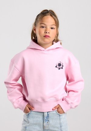 Jong meisje met lichtbruin haar, draagt een roze hoodie met een bloemenpatroon en lichtblauwe spijkerbroek met handen in de zakken.