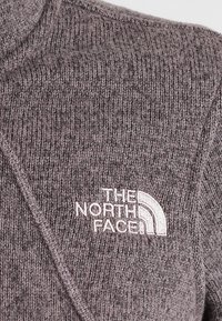 Primer plano de una tela de punto gris con el logotipo bordado en blanco "THE NORTH FACE" y un diseño curvado en una prenda.