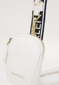 Vit läder crossbody-väska med en liten ficka, guldfärgade detaljer och ett texturerat band med svart text. Dragkedjedetalj och logotyp synlig.