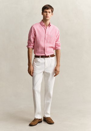 Junger Mann steht vor einfachem Hintergrund, trägt ein rosa-weiß kariertes Hemd, weiße Hose mit braunem Gürtel und braune Loafers.