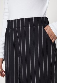 Schwarze Hose mit weißen vertikalen Streifen, ausgestattet mit einem breiten Bund und seitlichen Taschen. Der Stoff wirkt glatt mit einer feinen Struktur.