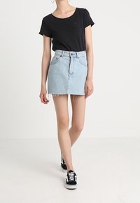 Frau in einem schwarzen Kurzarmoberteil, hellblauem Jeans-Minirock mit rohem Saum und schwarzen Turnschuhen, die vor einem weißen Hintergrund läuft.