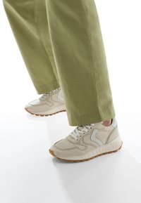 Sneaker beige in pelle traforata, con accenti bianchi e suola in gomma. Abbinato a pantaloni larghi verde oliva, che mettono in risalto le calzature.