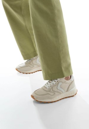 Beige sneaker with textured leather, white accents, and a rubber sole. Paired with loose-fitting olive green pants, emphasizing the footwear.
