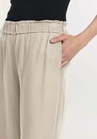 Beige linnen broek met een elastische tailleband, voorzien van zijzakken en een losse pasvorm. De stof heeft een gladde, zachte textuur.