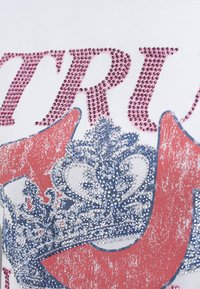 Motif décoratif de couronne et flèches avec strass roses et argentés sur tissu blanc, faisant partie d'un texte plus grand en strass roses.