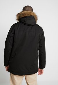 Černá parka s kapucí lemovanou kožešinou, se sametovým povrchem a delším střihem, zobrazená zezadu s béžovými kalhotami.