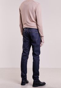 Pánské zúžené tmavě modré džíny s kontrastním oranžovým prošíváním, spárované se světle béžovou košilí s dlouhým rukávem a černými botami.