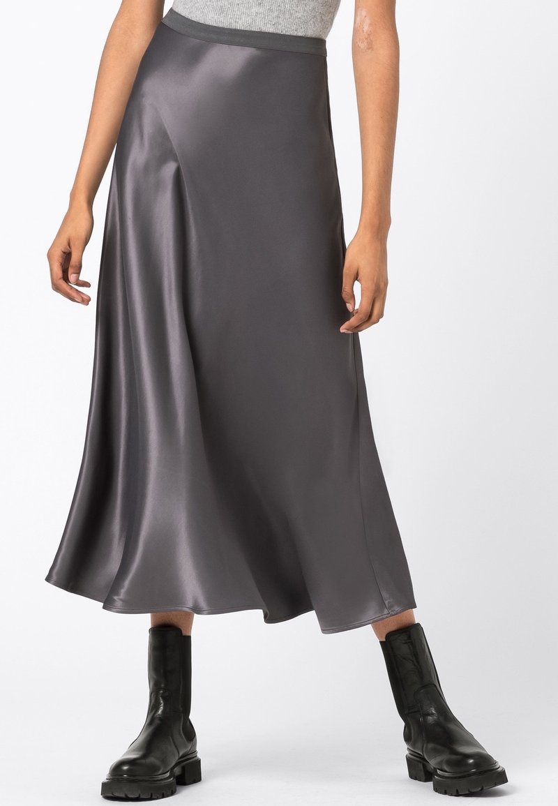 Femme portant une jupe midi en satin gris, un top sans manches gris et des bottines en cuir noires épaisses, debout devant un fond blanc.