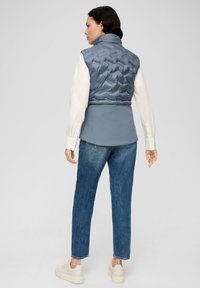 Gepolsterte blaue Weste mit hohem Kragen über einem grauen Oberteil, kombiniert mit blauen Jeans und hellen Sneakers, mit einem strukturierten, gesteppften Design.