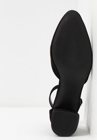 Chaussure à talon haut noire avec un bout pointu et des côtés ouverts, dotée d'une semelle texturée et d'une bride de cheville réglable. Matériau en daim doux.