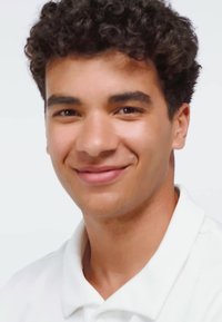 Młody mężczyzna o kręconych ciemnych włosach, uśmiechający się, ubrany w białą koszulę z kołnierzykiem, na jednolitym białym tle.