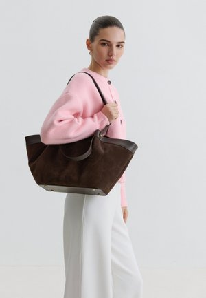 Femme en pull rose et pantalon blanc tenant un grand sac fourre-tout en cuir brun foncé sur l'épaule, face à la caméra avec une expression neutre.