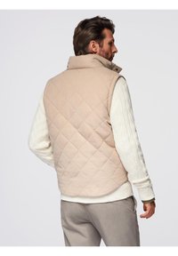 Gilet beige trapuntato con colletto alto, tasche laterali e una texture liscia, indossato sopra un maglione bianco lavorato a maglia a trecce.