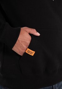Czarna bluza z kapturem z kieszenią kangurkiem, z pomarańczową metką "WRONG FRIENDS". Żebrowane mankiety i dół, gładka tekstura materiału.