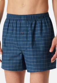 Blauwe geruite boxershort met een elastische tailleband, voorzien van een knoopsluiting aan de voorkant en een grijs ruitpatroon op een gladde stoftextuur.