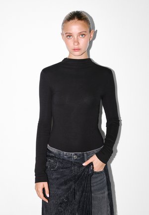 Fekete, hosszú ujjú felső, magas nyakkal. Simító, rugalmas anyagból készült. Szürke farmerrel párosítva, amely texturált derékkal rendelkezik.