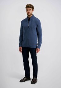 Blauer strukturierter Strickpullover mit halbem Reißverschluss, gerippten Bündchen und Saum. Kombiniert mit dunklen Hosen und dunklen Slippern.