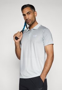 Pilkas polo marškinėliai su taškiniu raštu, trumpomis rankovėmis ir apykakle. Modelis laiko teniso raketę. Audinys atrodo lygus ir lengvas.