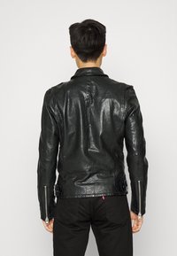 Młody mężczyzna odwrócony plecami, ubrany w czarną skórzaną kurtkę z zapięciem na suwak na rękawach i czarne spodnie, na jednolitym tle.