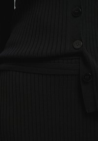 Indumento in maglia a coste nera con chiusura a bottoni e vita aderente. La texture è liscia con sottili linee verticali e bottoni a contrasto.