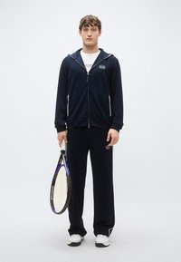 Chándal deportivo azul marino con detalles en blanco, que incluye una sudadera con capucha y cremallera, y pantalones. El modelo sostiene una raqueta de tenis y lleva zapatillas blancas.