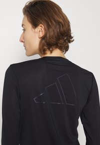 Crna dugorukava atletska majica s teksturiranom tkaninom, s istaknutim crnim logotipom na leđima, koja prikazuje elegantan moderan dizajn.