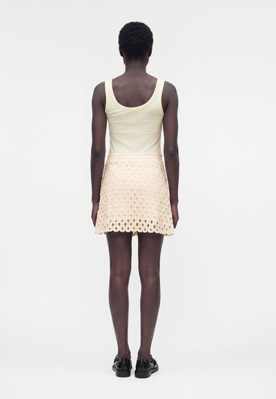 Άτομο που στέκεται πλάτη, φοράει αμάνικο λευκό τοπ, μπεζ φούστα με δαντέλα με τρύπες και μαύρα παπούτσια μπροστά σε μονόχρωμο φόντο.