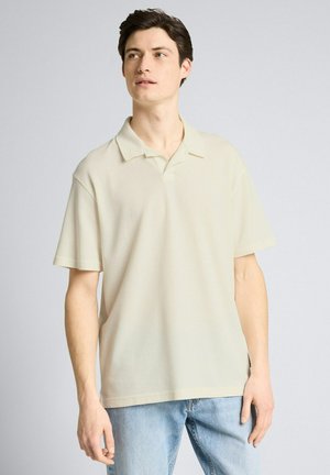 Junger Mann in cremefarbenem kurzärmligem Poloshirt und hellblauen Jeans, der leicht nach oben und zur Seite schaut.