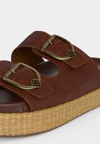 Καφέ δερμάτινο σανδάλι με δύο ρυθμιζόμενες ορείχαλκινες αγκράφες και πλεγμένη πλατφόρμα σε ανοιχτό φόντο.