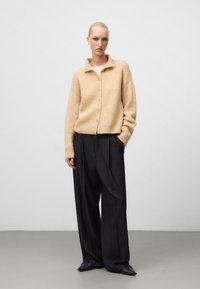 Cardigan en laine beige avec col et fermeture à bouton, associé à un pantalon noir taille haute à jambes larges et des chaussures noires à bout pointu.