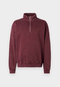 Pull-over en polaire bordeaux avec un col zippé mi-haut, des poignets côtelés et un petit logo brodé sur la poitrine. Texture douce et design décontracté.