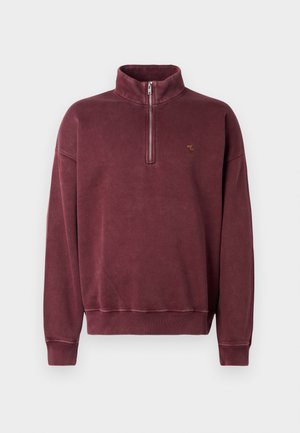 Burgundy fleece pullover with a half-zip collar, ribbed cuffs, and a small embroidered logo on the chest. Soft texture and casual design.