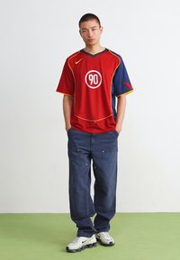 Rød t-shirt med korte ærmer, blå detaljer og gule kanter, udstyret med et cirkulært hvidt grafisk motiv og løse, mørkeblå jeans med lommer.