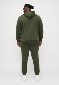 Olivgrünes Sweatshirt und Jogginganzug-Set aus weichem Stoff, mit Kapuze, gerippten Bündchen und schmal zulaufenden Beine, kombiniert mit weißen Sneakers.