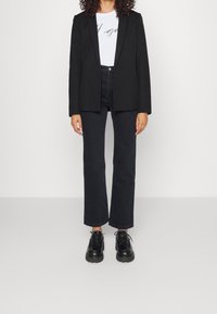 Svart skräddarsydd kavaj över en vit skjorta med grå text, tillsammans med mörka jeans och kraftiga svarta snörskor. Slät tyg och strukturerad passform.