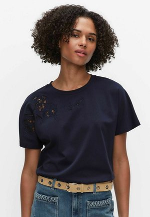 Kvinde med krøllet hår iført en mørk navyblå top med blomsterudskæringsbroderi på skulderen, stoppet ned i blå jeans med beige bælte.