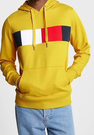 Mężczyzna w żółtej bluzie z kapturem z poziomymi blokami kolorów: czarnym, białym, czerwonym i granatowym, w jasnoniebieskich dżinsach, z rękami w kieszeniach bluzy.