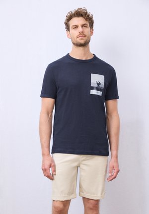 T-shirt bleu marine à manches courtes en tissu doux avec un détail de poche graphique rectangulaire blanche présentant un design de silhouette.
