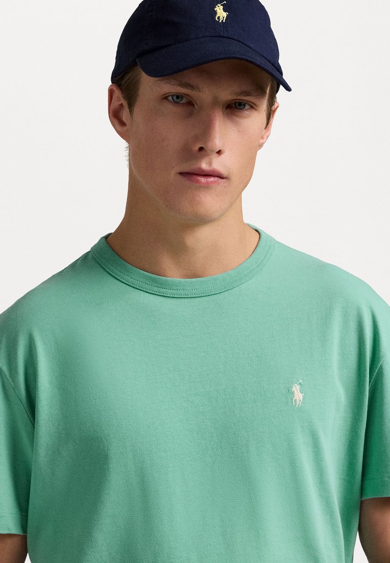 Mintgrünes Baumwoll-T-Shirt mit Rundhalsausschnitt und kurzen Ärmeln, mit einem kleinen gestickten Logo auf der linken Brust. Dunkelblappe Kappe.