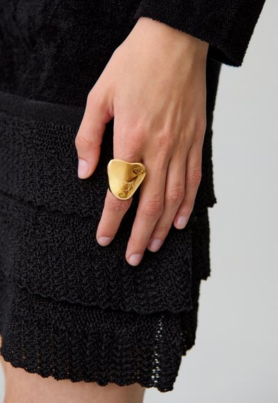 Anillo de oro con un diseño suave en forma de corazón y detalles grabados, llevado en una mano junto a una prenda de tejido negro texturizado.