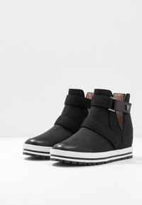 Zwarte, gestructureerde enkel sneakers met dikke witte zolen en brede stoffen banden die over de voorkant en rond de enkel kruisen.