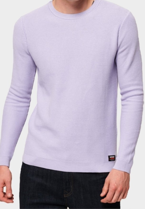 Homme portant un pull à manches longues violet clair avec col rond, associé à un pantalon foncé, se tenant devant un fond uni.